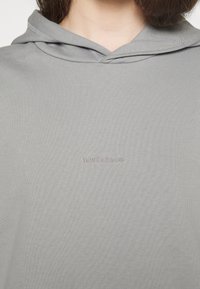 Felpa grigia chiaro realizzata in morbido tessuto, dotata di cappuccio con coulisse e di un piccolo logo "new balance" rosa pallido nella zona del petto.