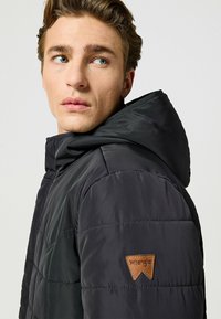 Giacca a vento nera con cappuccio, caratterizzata da una texture trapuntata e una toppa in pelle beige con il logo Wrangler sulla manica sinistra.