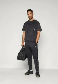 Svart falmet T-skjorte med en liten logo, kombinert med svarte joggebukser og svarte joggesko. Holder en svart duffelbag.