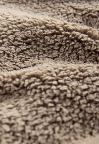 Tissu beige texturé avec un poil doux et en relief. La surface semble chaude et douce, présentant des motifs irréguliers et ondulés sur toute la surface.