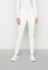 Mujer llevando jeans ajustados blancos, zapatillas blancas con motas de colores y una camiseta de manga larga blanca acanalada, de pie contra un fondo liso.
