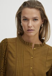 Blusa di cotone marrone con scollatura arricciata, bottoni, pieghe verticali e maniche a palloncino ricamate con motivi ad occhio di pernice.