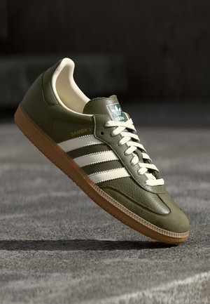 Buty sportowe Adidas Samba w oliwkowej zieleni z białymi paskami, beżowym wnętrzem i gumową podeszwą unoszące się nad teksturowaną szarą powierzchnią.