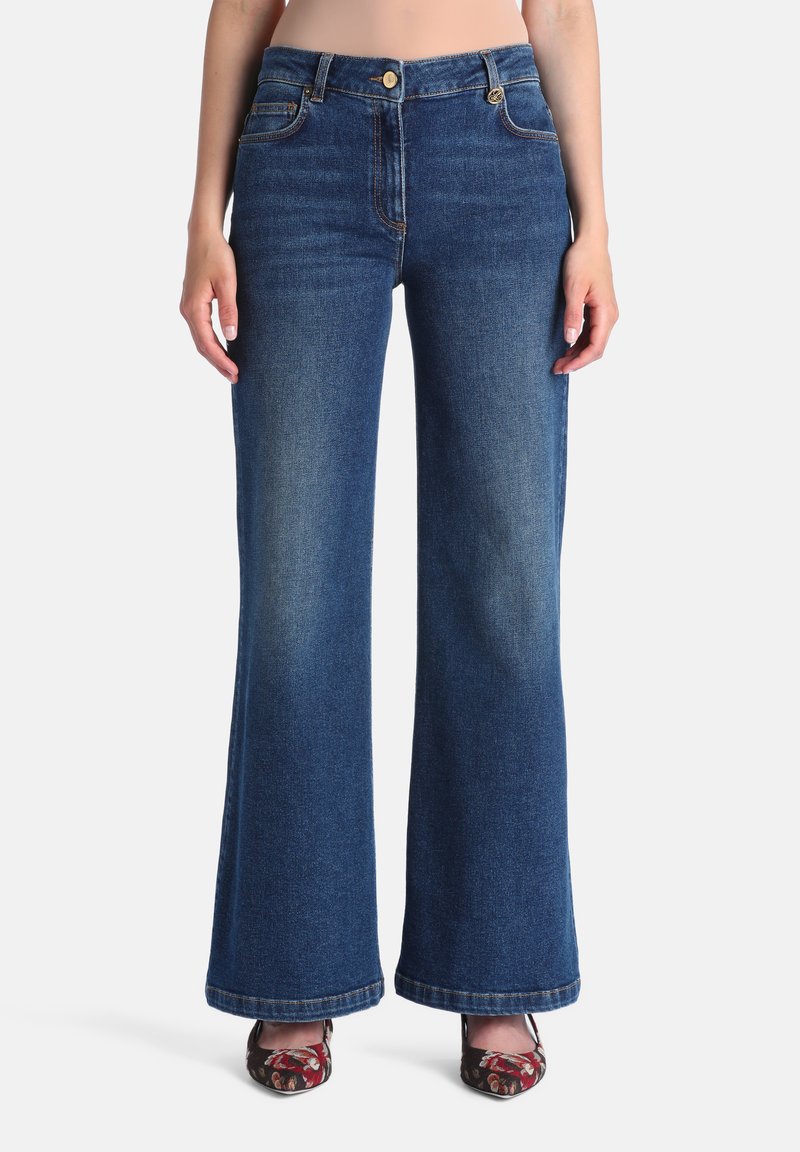 Femme portant un jean flare bleu foncé taille haute avec fermeture bouton et zip, associé à des talons pointus à motif floral, debout sur fond blanc.