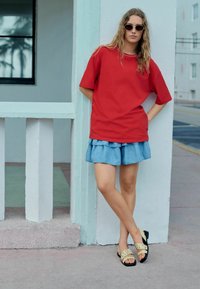 Rotes übergroßes T-Shirt, geschichteter Jeansrock und tanfarbene Rutschsandalen mit Ausstanzungen. Der Hintergrund zeigt eine hellblaue Wand und ein Fenster.
