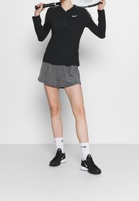 Zwarte long sleeve top met rits, grijze shorts, witte sokken en zwarte sportschoenen. Inclusief witte Nike logo accenten en een tennisracket.