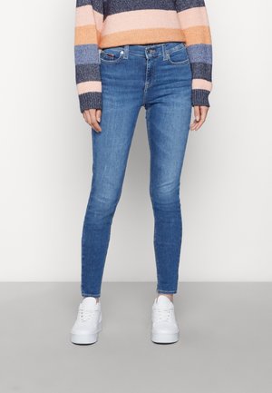 Personne portant un jean skinny bleu, un pull rayé multicolore et des baskets blanches sur un fond uni.