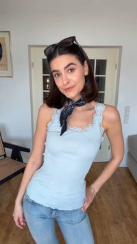 Γυναίκα με κοντά σκούρα μαλλιά, φορά γυαλιά ηλίου στο κεφάλι, ανοιχτό μπλε τοπ με δαντέλα, μπλε μαντήλι γύρω από το λαιμό και ανοιχτό μπλε τζιν, σε ένα δωμάτιο.