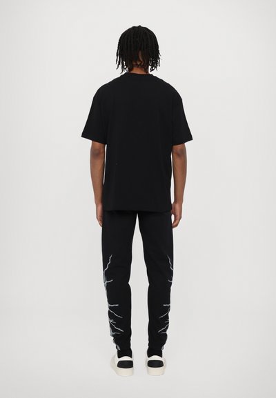 T-shirt in cotone nero con una vestibilità rilassata, abbinato a pantaloni della tuta neri con design di fulmini bianchi lungo i lati.