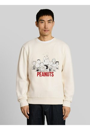 Man draagt een crèmekleurige trui met zwart-witte Peanuts-figuren en rode "PEANUTS" tekst aan de voorkant, staand tegen een grijze achtergrond.