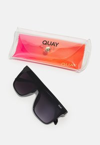 Czarne, oversize prostokątne okulary przeciwsłoneczne z ciemnymi szkłami i logo Quay obok przezroczystego plastikowego etui z różowo-pomarańczową ściereczką Quay w środku.
