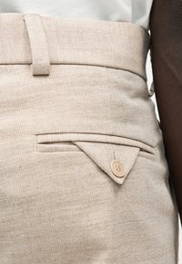 Κοντινό πλάνο σε μπεζ παντελόνια με σχέδιο ψαροκόκαλο, με ζώνη στη μέση, θηλιά για ζώνη, πλαϊνή τσέπη και λεπτομέρεια με κουμπωτό καπάκι.