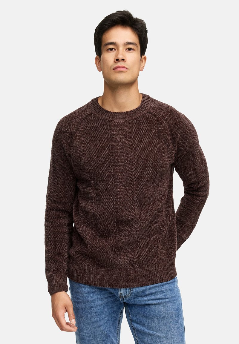 Jonge man met kort, donker haar, gekleed in een gestructureerde donkerbruine trui met ronde hals en blauwe jeans, staand met één hand achter zijn rug.