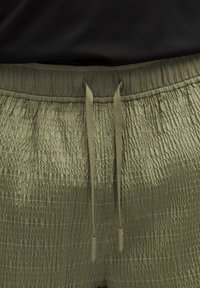 Olīvu zaļas bikses ar teksturētu apdari, ar elastīgu jostu un saistēm. Audumam ir smalka režģa raksts visā virsmā.
