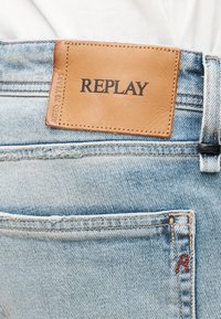 Φωτεινά μπλε τζιν από denim με ένα καφέ δερμάτινο ταμπελάκι με την ένδειξη "REPLAY" και ένα μικρό ραμμένο κόκκινο "R" στην πίσω τσέπη.