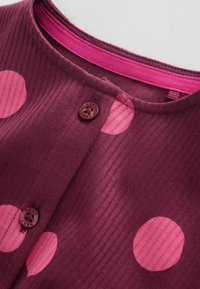 Gros plan sur un tissu violet côtelé avec de gros pois roses, col rond avec bordure rose et deux boutons violets étiquetés "NEXT".