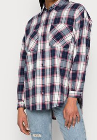 Camicia di flanella a quadri blu navy e bianca con accenti rossi, design con bottoni, due tasche sul petto, maniche lunghe e vestibilità rilassata.