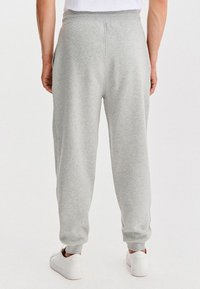 Mężczyzna ubrany w jasnoszare dresowe spodnie i białe sneakersy, stoi tyłem na tle białego tła.