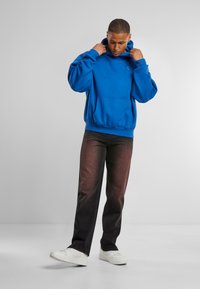 Modra kapuča s prednjim žepom, v kombinaciji s temnimi gradientnimi jeans hlačami in belimi športnimi copati. Figura stoji na nevtralni podlagi.