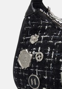 Sac à main en tweed noir et blanc avec quincaillerie argentée, comportant plusieurs badges décoratifs et une fermeture éclair, complété par un tissu texturé.