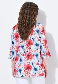 Blommig tunika i skjortstil med röda, blå och vita blommor på en ljus bakgrund, med trekvartslånga ärmar och en lös passform.