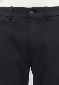 Nahaufnahme des Taillenbands schwarzer Hosen mit Gürtelschlaufen und einem Knopfverschluss vor weißem Hintergrund.