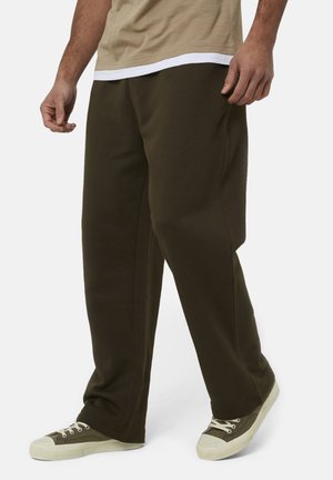 Homme portant un pantalon large kaki foncé et des baskets basses assorties avec des semelles blanches et des lacets, sur un fond blanc.