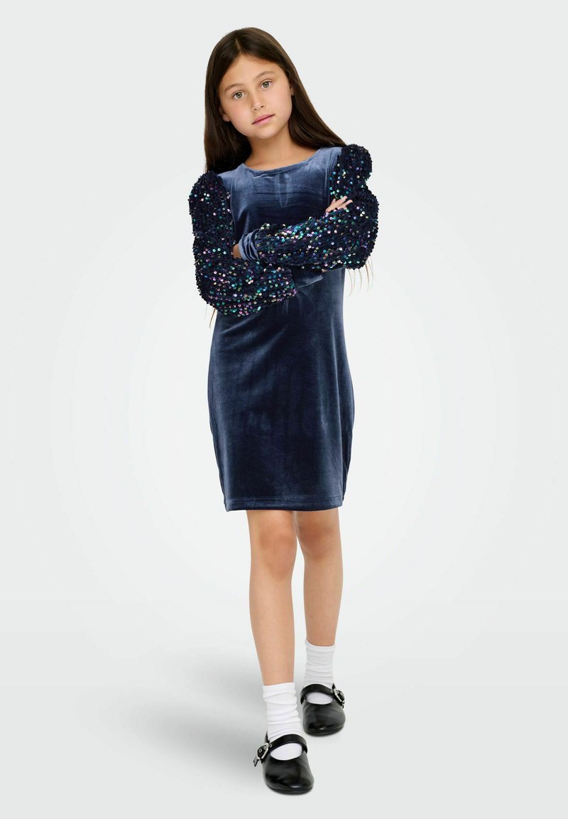 Robe en velours bleu marine avec des manches bouffantes ornées de sequins, arborant un col rond et une coupe au genou, associée à des chaussures noires et des chaussettes blanches.