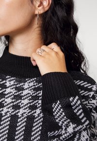 Pull en tricot noir et blanc avec un col côtelé haut, arborant un motif pied-de-poule. Le modèle porte des boucles d'oreilles en argent et des bagues.