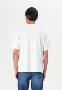 Balta trumpa rankovė t-shirt su apvaliu kaklu, pagaminta iš lygaus medvilnės. Nugaroje, derinama su mėlynomis džinsinėmis kelnėmis. Minimalistinis dizainas, be matomų raštų.