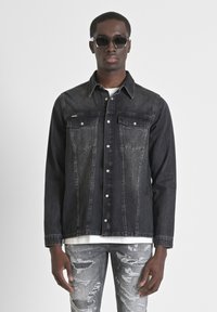 Chaqueta de mezclilla negra con dos bolsillos en el pecho y cierre de botones. Usada sobre una camiseta blanca, combinada con jeans grises desgastados. Textura suave.