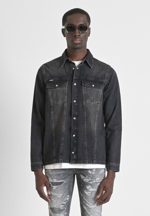 Giacca di jeans nera con due tasche sul petto e chiusura a pulsante. Indossata sopra una camicia bianca, abbinata a jeans grigi strappati. Tessuto liscio.