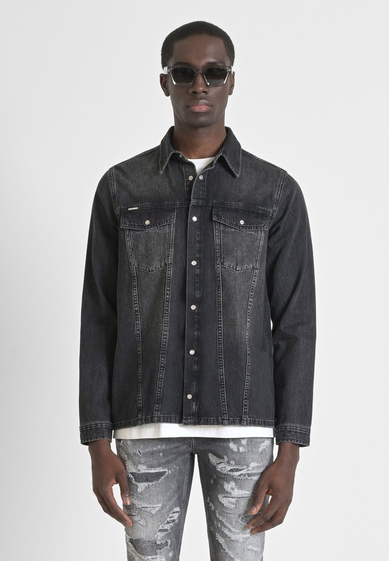 Chaqueta de mezclilla negra con dos bolsillos en el pecho y cierre de botones. Usada sobre una camiseta blanca, combinada con jeans grises desgastados. Textura suave.