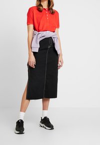 Femme portant un polo rouge à manches courtes, une jupe midi noire avec des coutures blanches et une fente sur le côté, des baskets noires avec des semelles blanches, et un sweatshirt lavande noué à la taille.