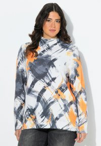 Frau mit welligem, dunklem Haar, die einen weißen Rollkragenpullover mit abstrakten schwarzen und orangefarbenen Pinselstrichmustern sowie dunklen Hosen trägt.