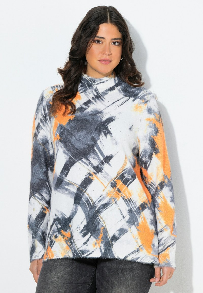 Frau mit welligem, dunklem Haar, die einen weißen Rollkragenpullover mit abstrakten schwarzen und orangefarbenen Pinselstrichmustern sowie dunklen Hosen trägt.