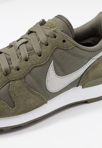 Olive green Nike sneaker with suede and mesh, white swoosh logo, lace-up closure, and white midsole.