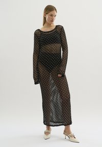 Rochie din plasă neagră cu mâneci lungi, prezentând buline albe mari, un design transparent și o tăietură până la podea. Asortată cu pantofi cu toc bej.