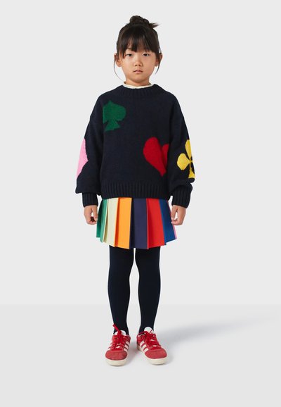 Marineblauer Pullover mit bunten Herz-, Pik- und Blumenmustern; mehrfarbiger Faltenrock; schwarze Strumpfhosen; rote Sneakers mit weißen Streifen.