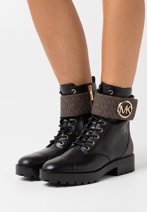 Bottes chelsea en cuir noir avec des lacets, ornées de larges revers marron épais, d'une fermeture éclair dorée et d'un logo rond doré MK sur le côté.