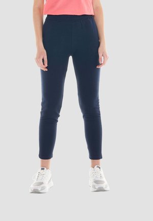 Femme portant un pantalon slim bleu marine, un haut corail et des baskets blanches, debout devant un fond uni clair.