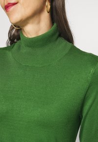 Vihreä pooloneule, jossa on istuva malli ja resorikuvioinen pinta. Kangas näyttää sileältä ja joustavalta, korostaen tiivistä ja imartelevaa istuvuutta.