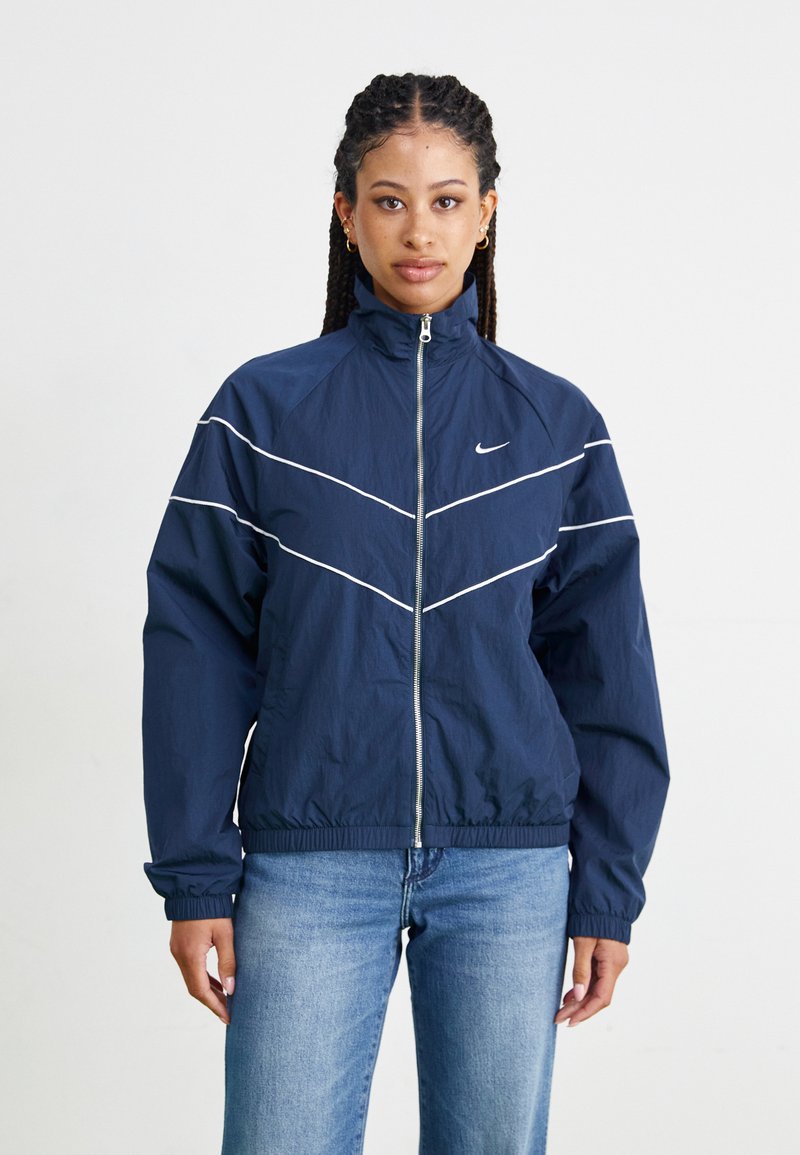 Veste coupe-vent bleu marine avec un col montant, des fermetures éclair argentées et des rayures en chevrons blanches sur la poitrine et les manches.