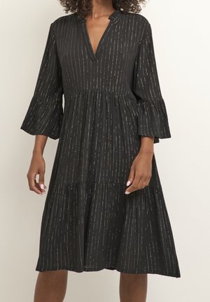 Femme portant une robe midi noire avec des rayures verticales argentées, des manches cloche et un décolleté en V sur un fond clair uni.