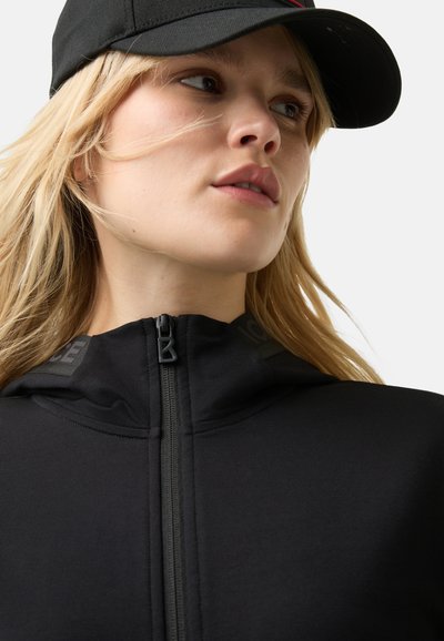 Schwarze Zip-Hoodie mit hohem Kragen, strukturiertem Stoff, Logo-Akzenten auf den Schultern und einem frontalen Reißverschluss. Getragen mit einer schwarzen Mütze.