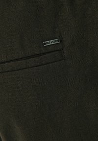 Teléfono de textura verde oscuro con un patrón sutil, que presenta un pequeño acento de logo metálico en la costura lateral y un detalle de bolsillo.