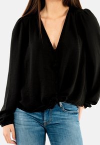 Femme portant un blouse noire à manches longues en V avec des boutons et un détail de nœud, associée à un jean en denim bleu, se tenant avec une main dans la poche.