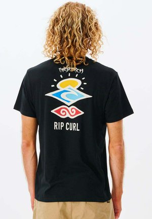 Person mit lockigem blondem Haar trägt ein schwarzes Rip Curl T-Shirt mit Sonne-, Wellen- und rotem Spiralmotiv auf der Rückseite, kombiniert mit beigen Shorts.