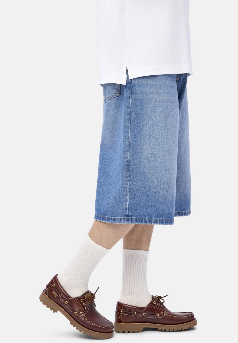 Personne portant un short en denim léger bleu ciel, s'arrêtant aux genoux, avec des chaussettes blanches et des chaussures bateau en cuir marron sur fond blanc.
