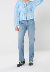 Světle modrá blůza s balónovými rukávy a detaily na šňůrku, spárovaná se světle modrými džíny rovného střihu a černými teniskami se zelenými tkaničkami.